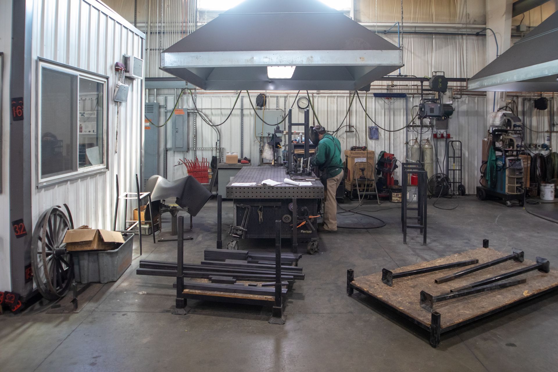Welding workshop interior with a person working at a metal table under a large hood. Metal pieces are nearby.
