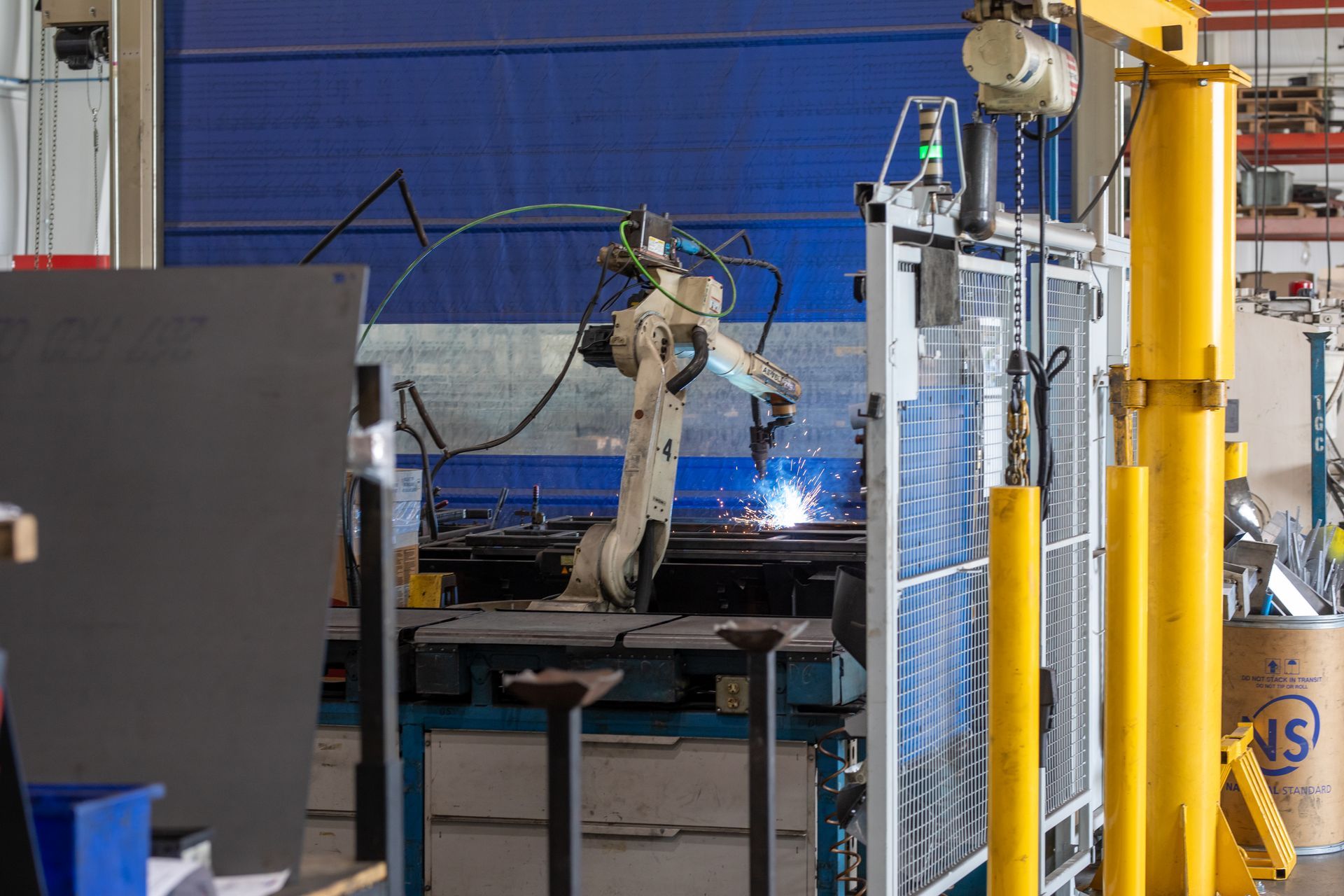 Robot welding metal in a factory setting. Blue, white, and yellow machinery. Sparks and safety fencing present.
