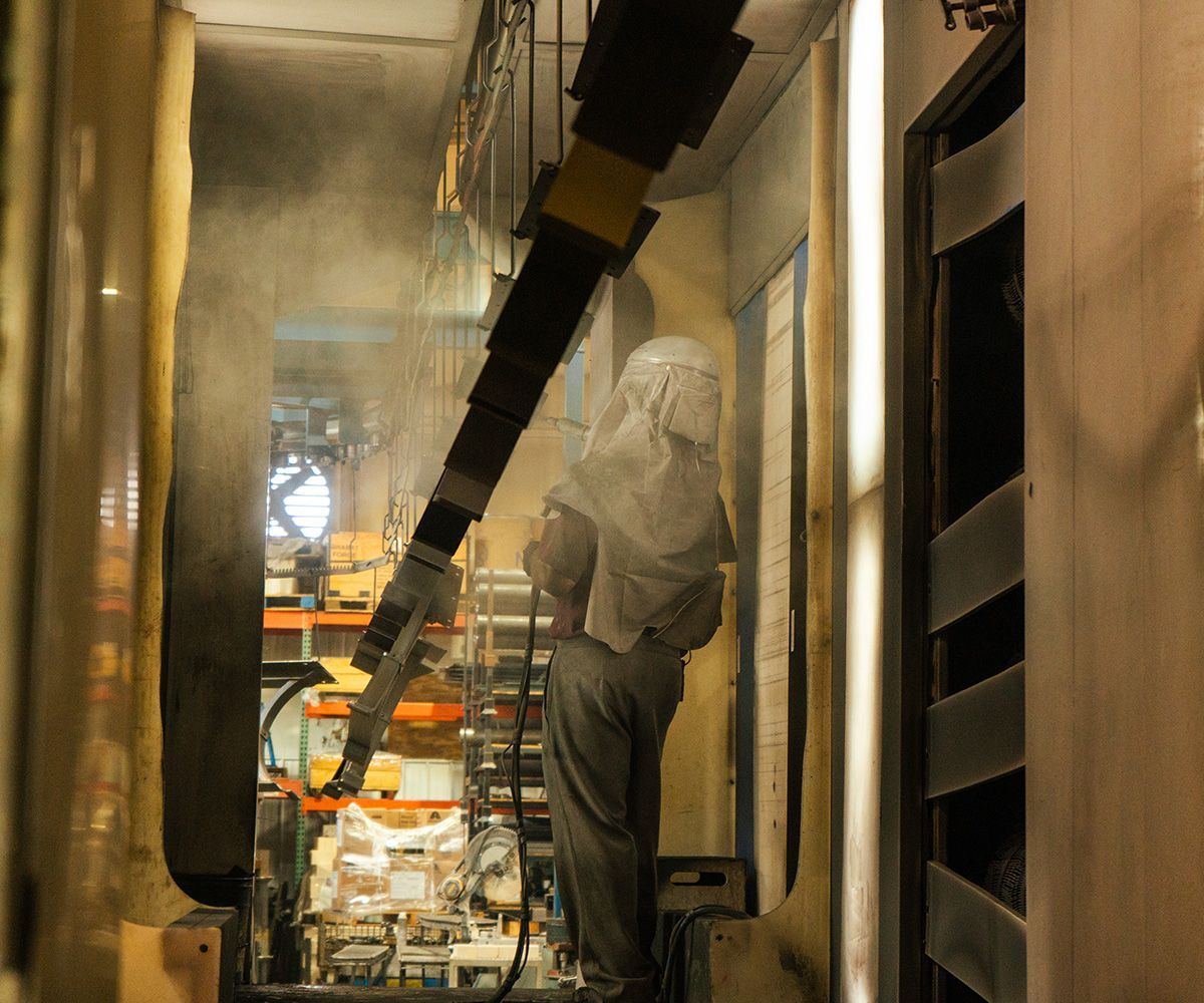 Person in protective gear sprays paint in a factory, with hanging metal parts and equipment visible.
