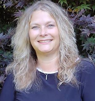 Smiling woman with wavy blonde hair in a dark top, standing in front of leafy greenery.
