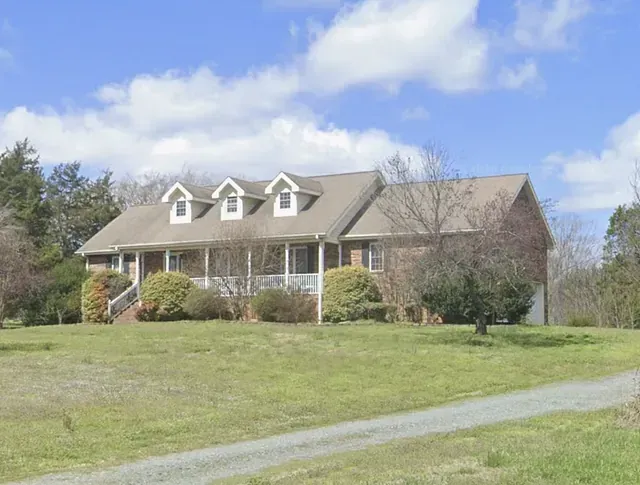 Brick house with dormer windows and a front porch on a grassy hill under a blue sky.