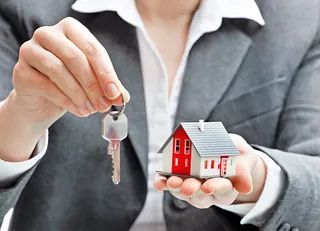 Person holding house keys above a small model house in their palm