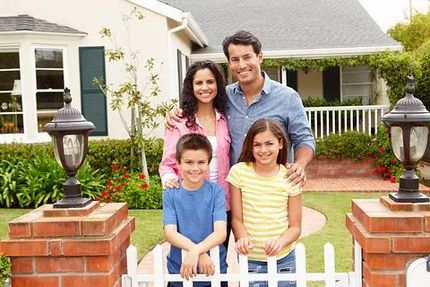 Family standing behind a white picket fence in front of a house, smiling in a yard with brick pillars