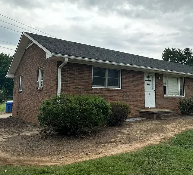 Brick house with white trim, front door, windows, and shrubs under a cloudy sky.