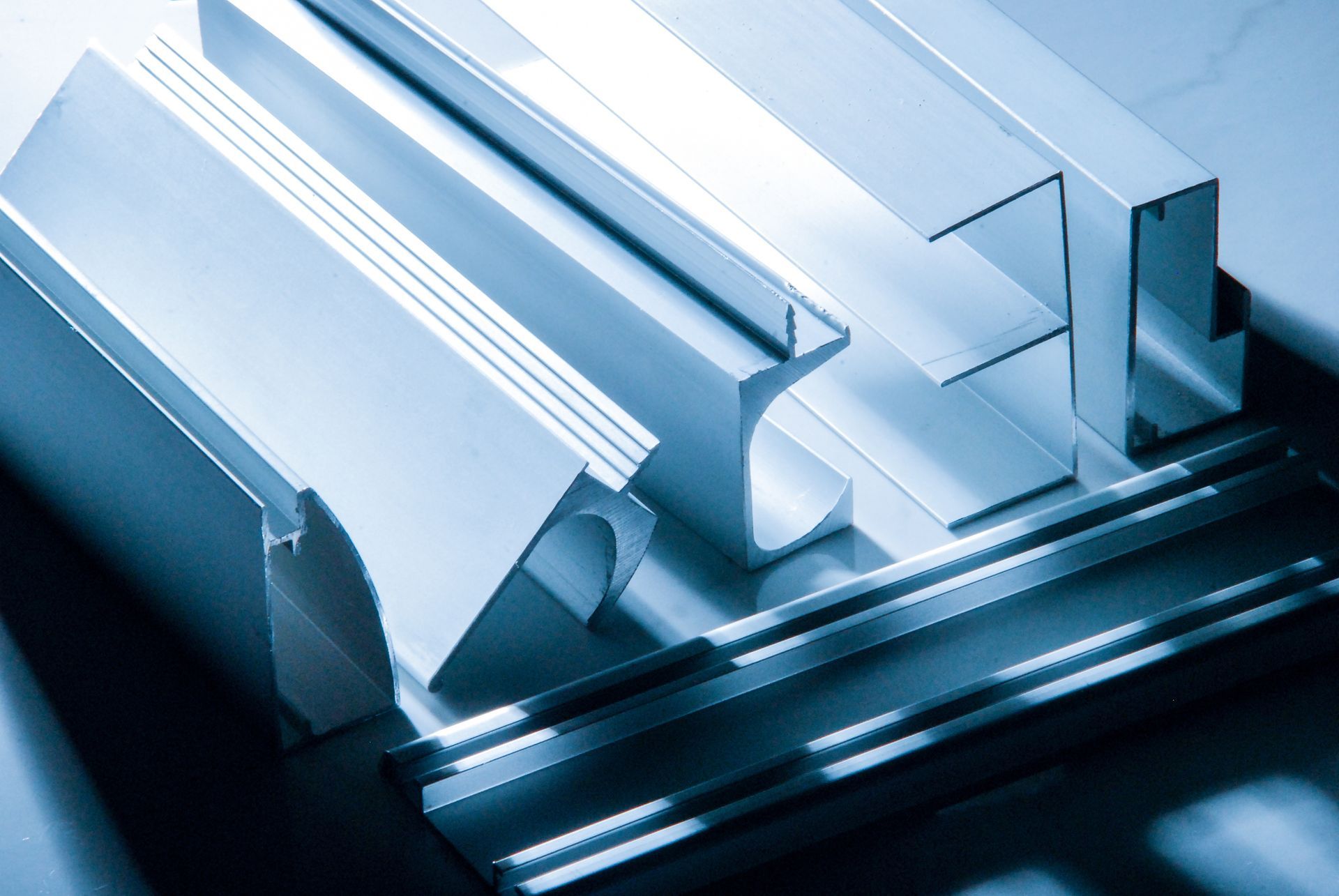 Assortiti profili ed estrusi in alluminio metallico disposti su una superficie scura sotto una luce fredda dai toni bluastri.