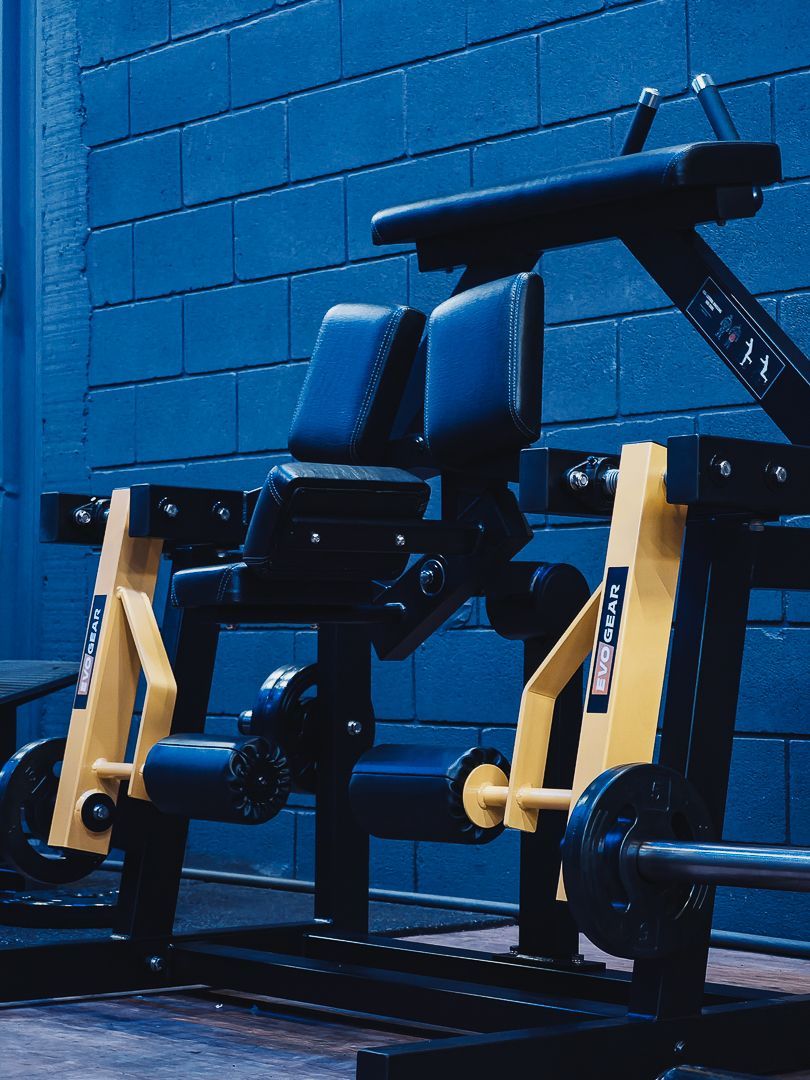Máquina de leg press azul e preta em uma academia, encostada em uma parede de tijolos azuis.