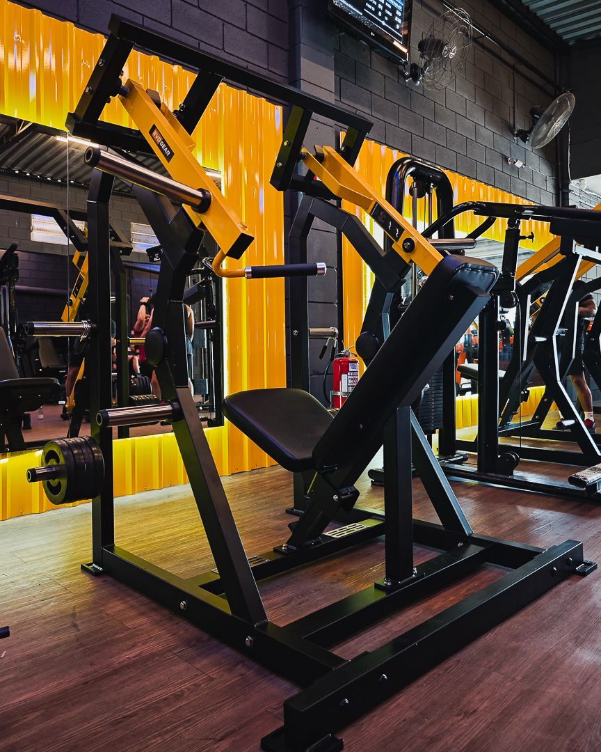 Aparelhos de musculação amarelos e pretos em uma academia com espelhos e piso de madeira.