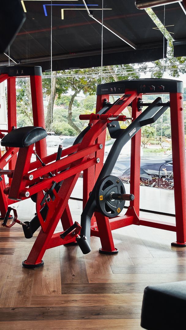 Aparelho de musculação vermelho e preto ao lado de grandes janelas em um estúdio de fitness iluminado.