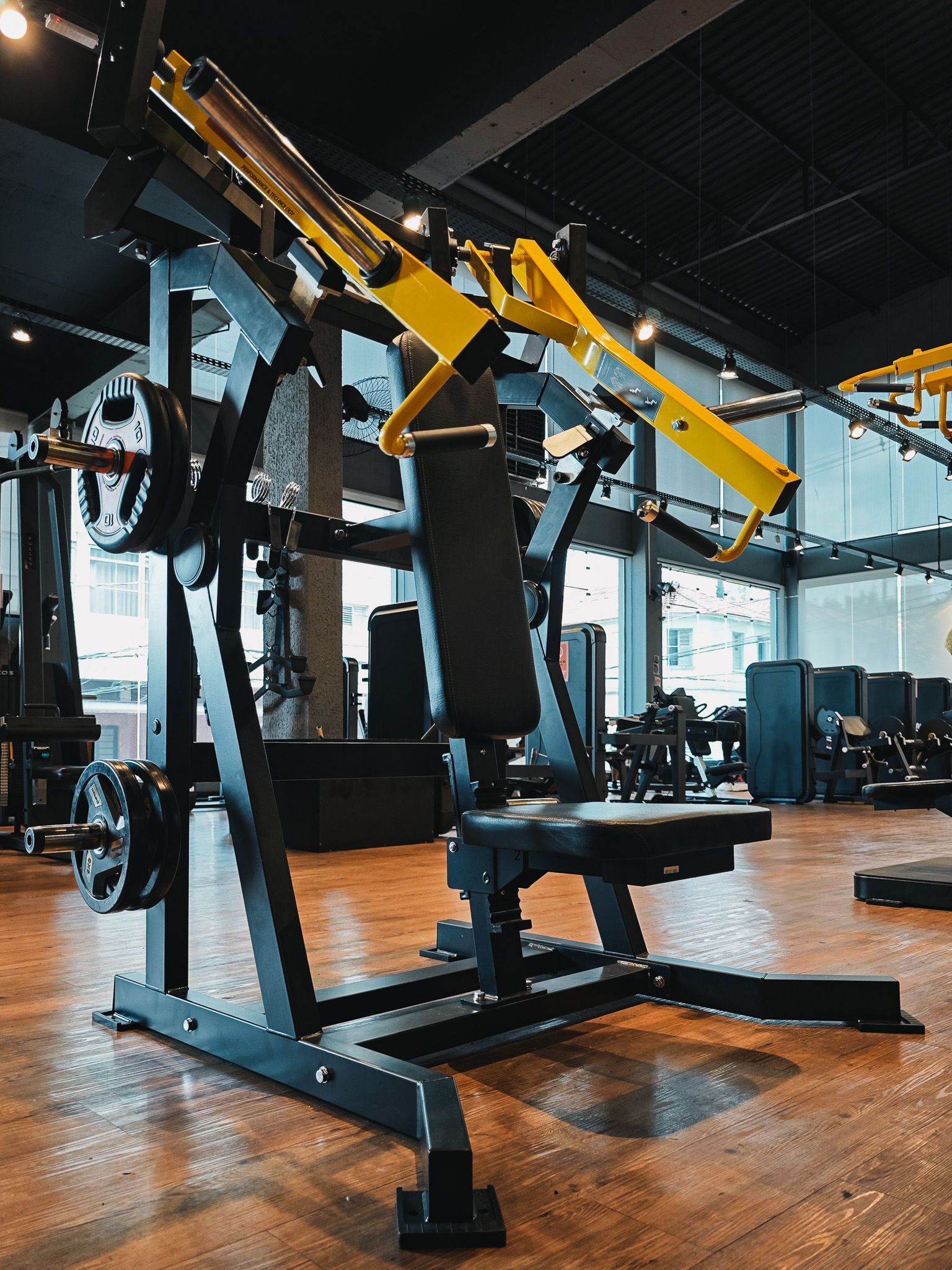 Aparelhos de musculação amarelos e pretos em uma academia moderna com piso de madeira e janelas amplas.
