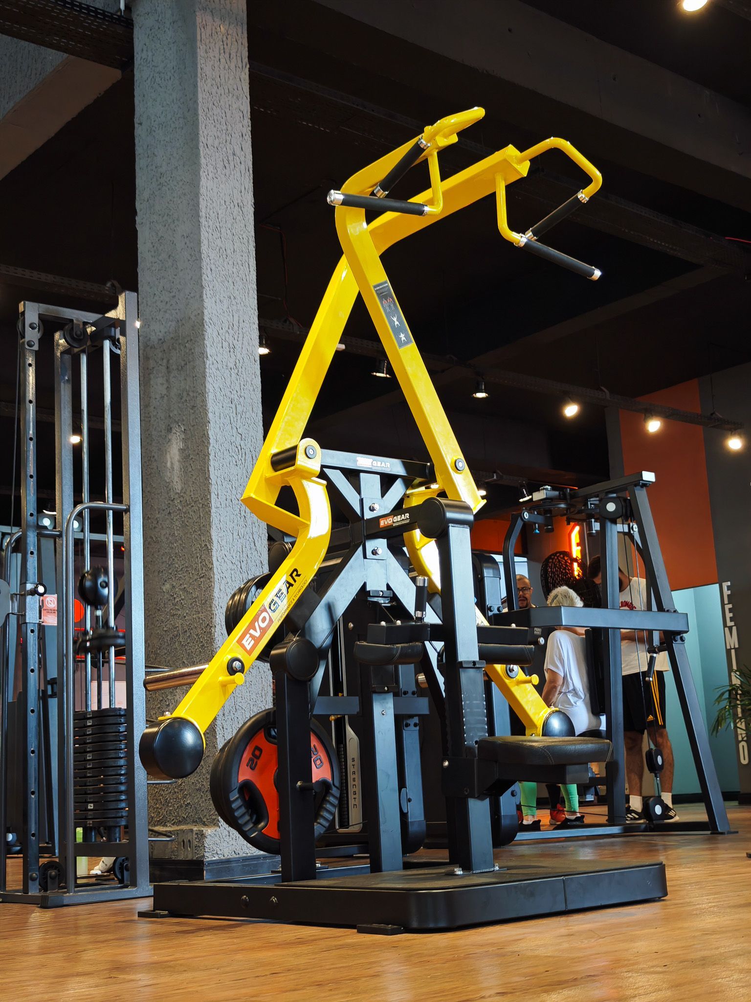 Aparelho de musculação amarelo em uma academia com outros equipamentos de ginástica ao fundo.