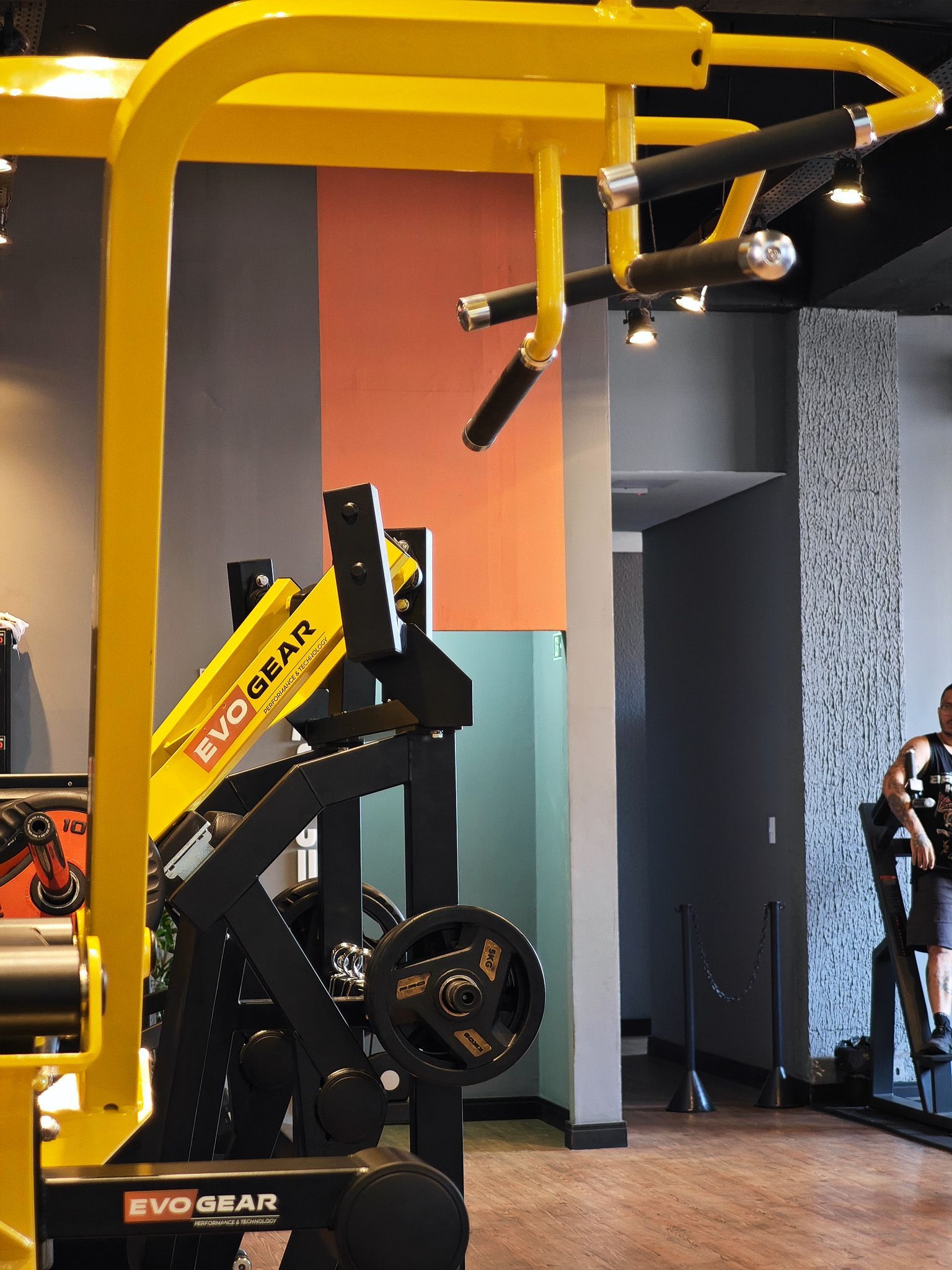 Aparelho de ginástica amarelo Evo Gear em uma academia com espelhos e equipamentos pretos.