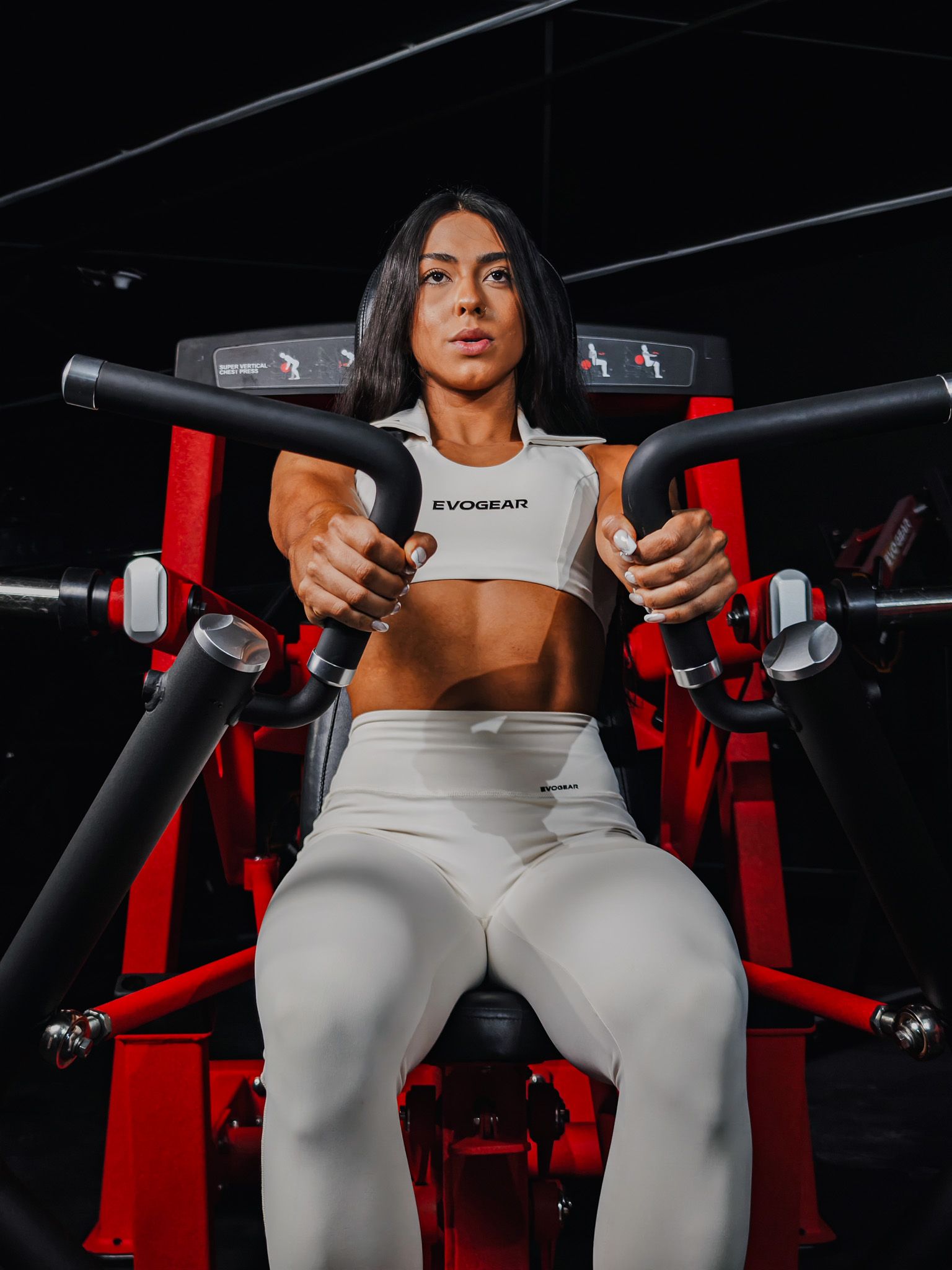 Mulher sentada em um aparelho de ginástica vermelho, segurando barras pretas contra um fundo escuro de estúdio.