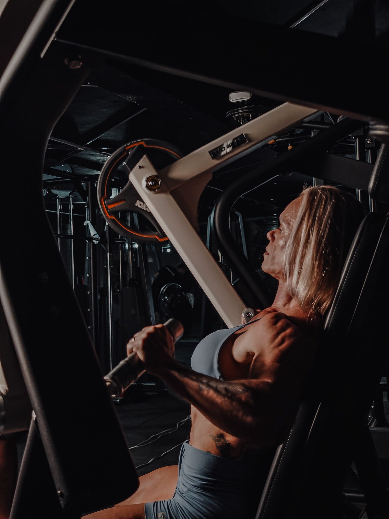 Mulher se exercitando em uma máquina de cabos em uma academia pouco iluminada, segurando as alças enquanto está sentada.