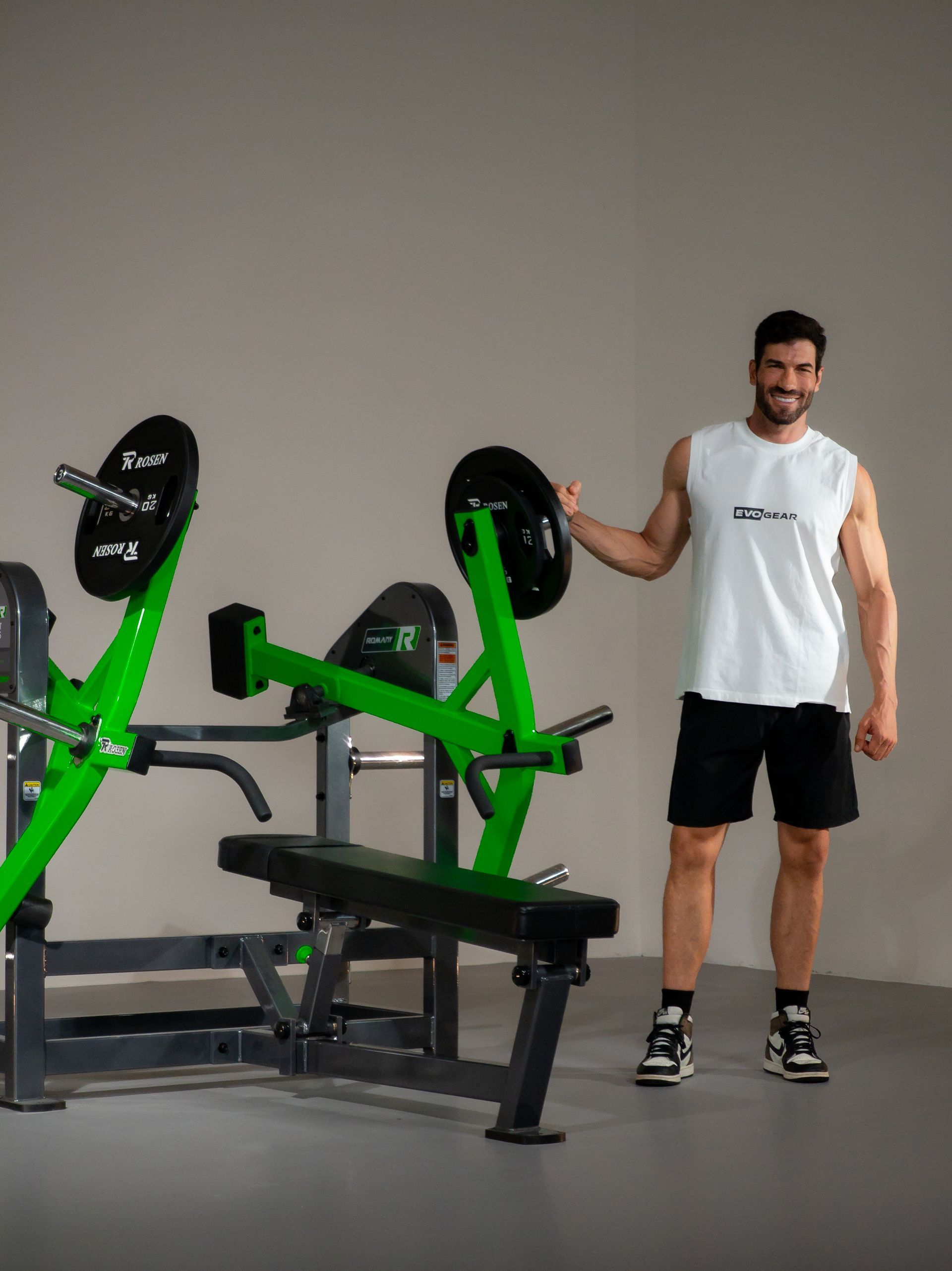 Homem posando ao lado de um aparelho de musculação verde e preto em um estúdio de ginástica.
