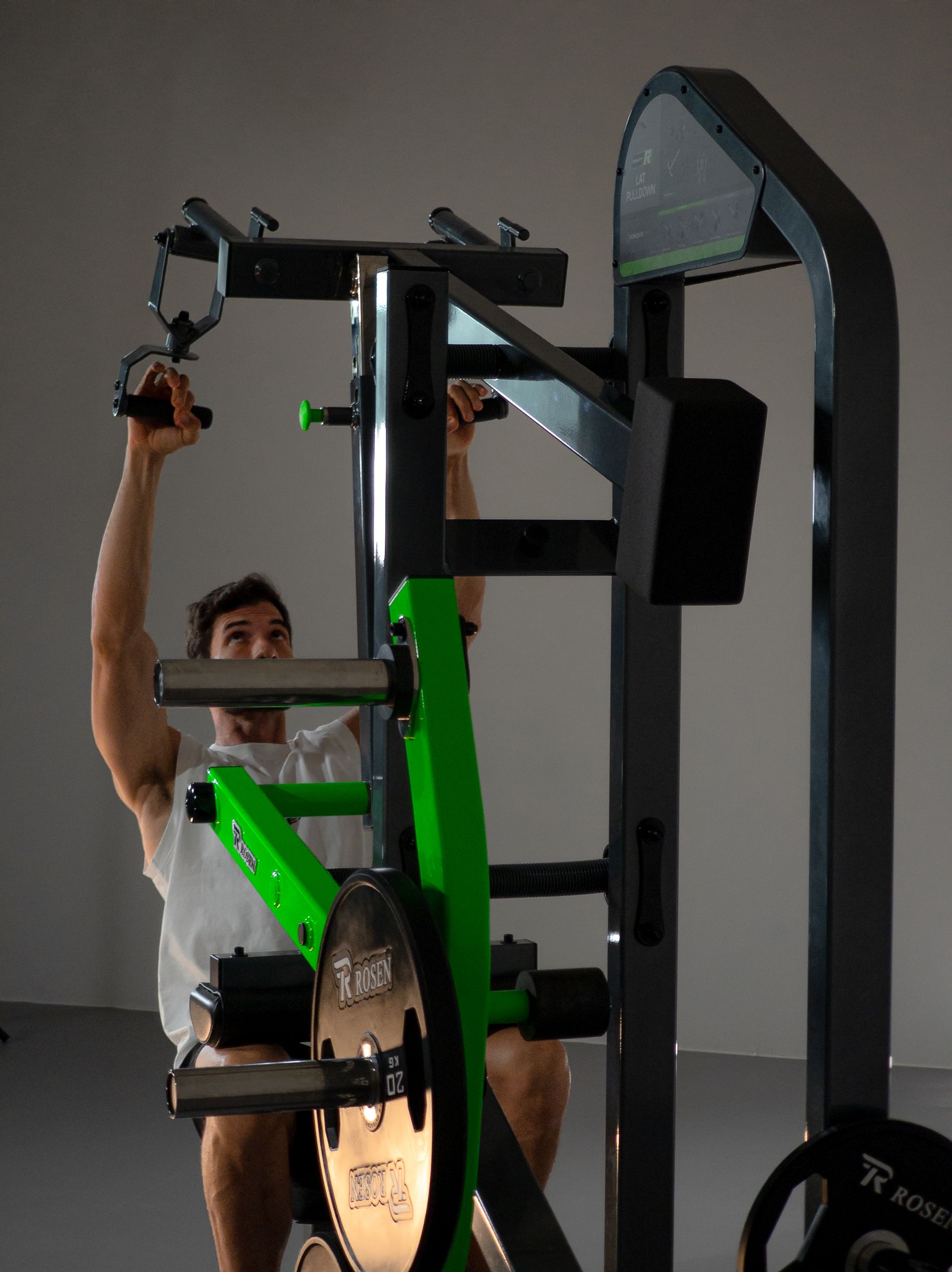 Pessoa utilizando um aparelho de ginástica verde e preto, segurando as barras superiores em uma sala de musculação.