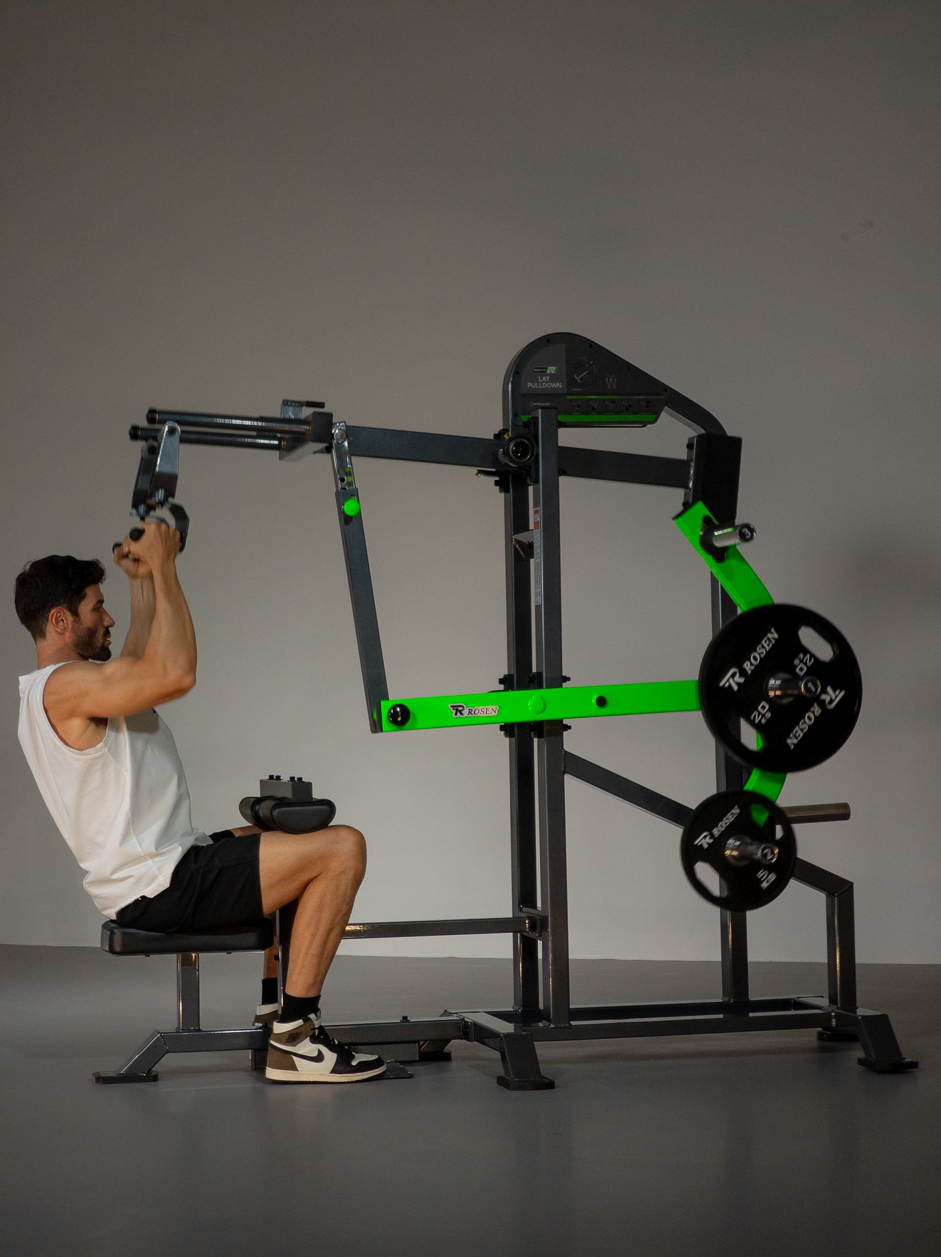 Homem usando um aparelho de musculação preto e verde em uma academia, sentado e puxando uma alavanca acima da cabeça.