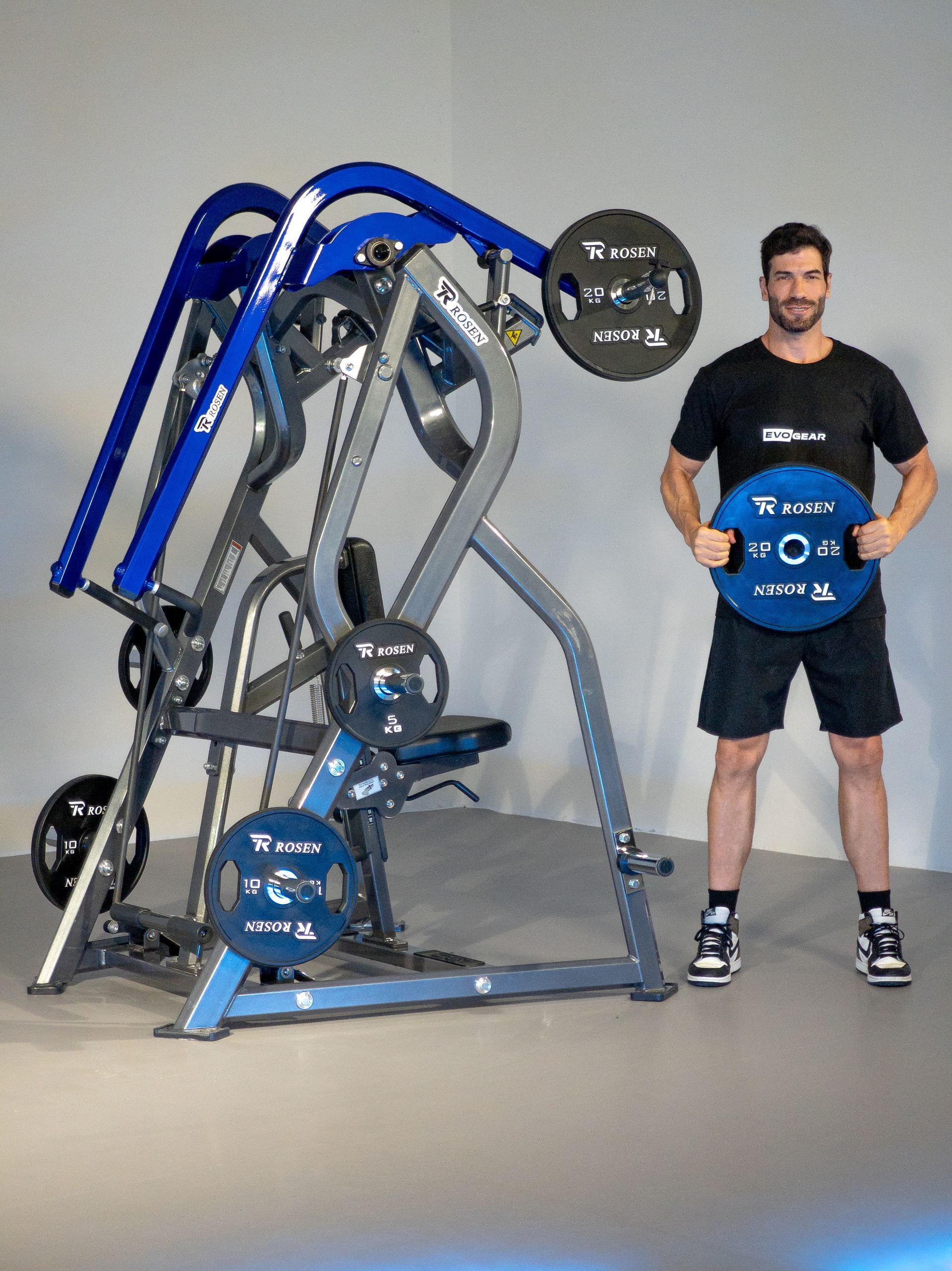 Homem em pé ao lado de um aparelho de musculação azul e cinza em uma academia, segurando uma anilha azul.