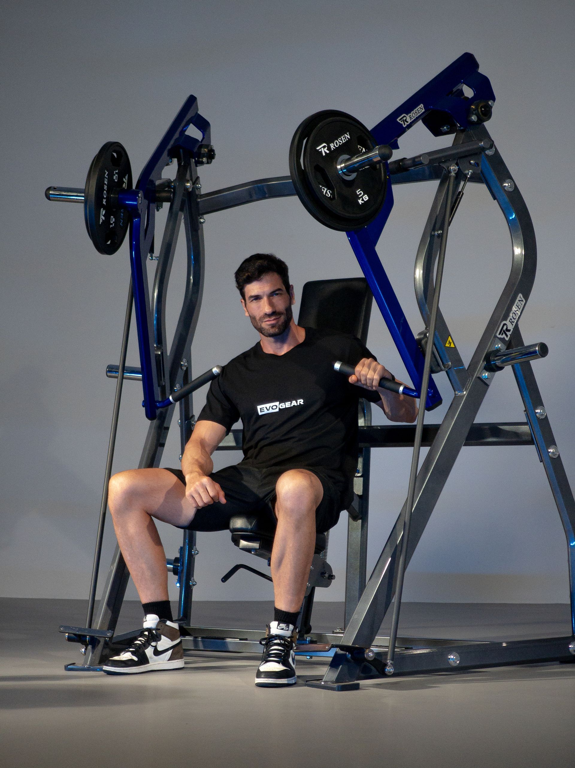 Homem sentado em uma máquina de leg press em uma academia, com equipamentos azuis e prateados.