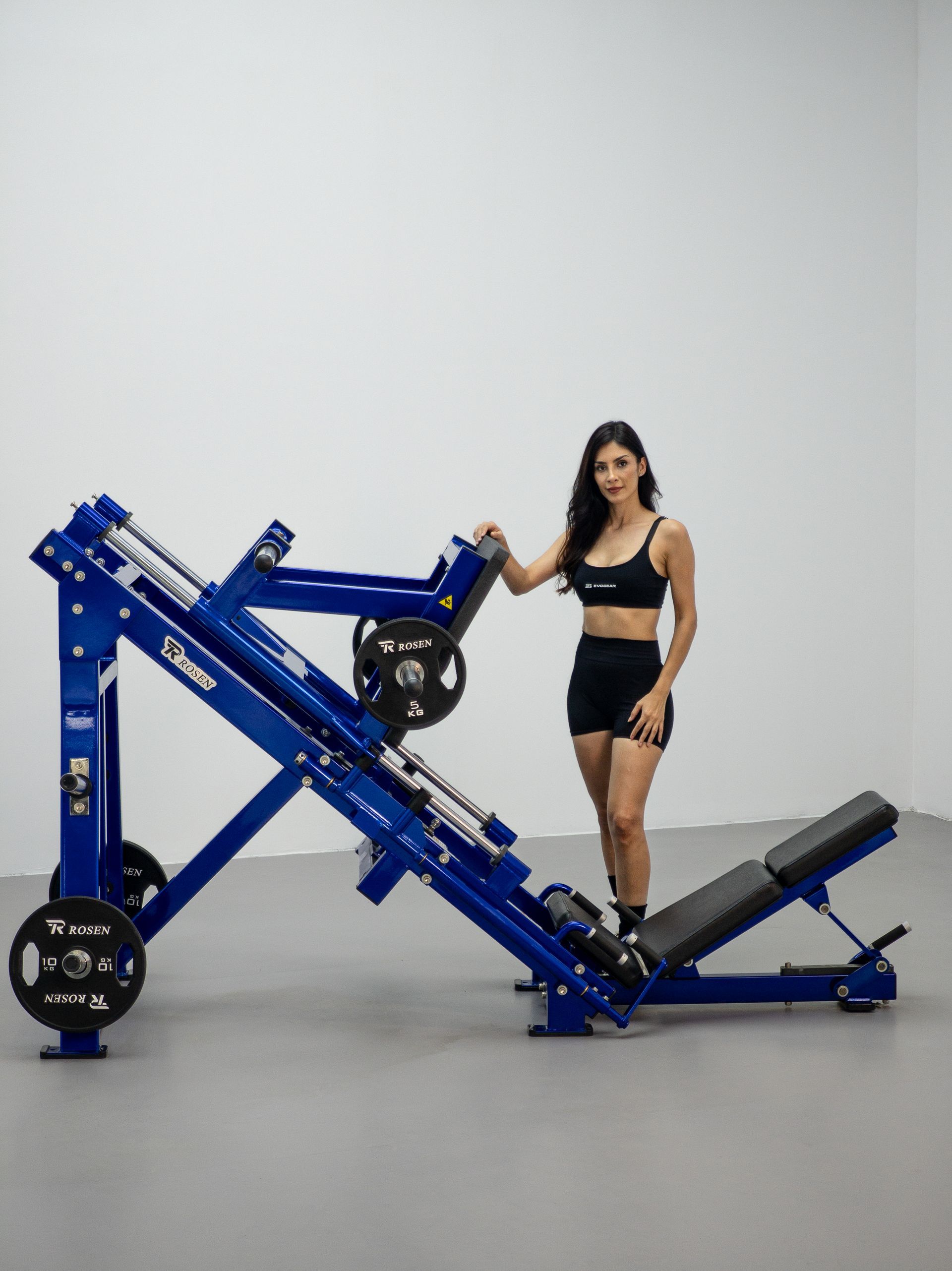 Mulher em pé ao lado de uma máquina de leg press azul em uma academia.
