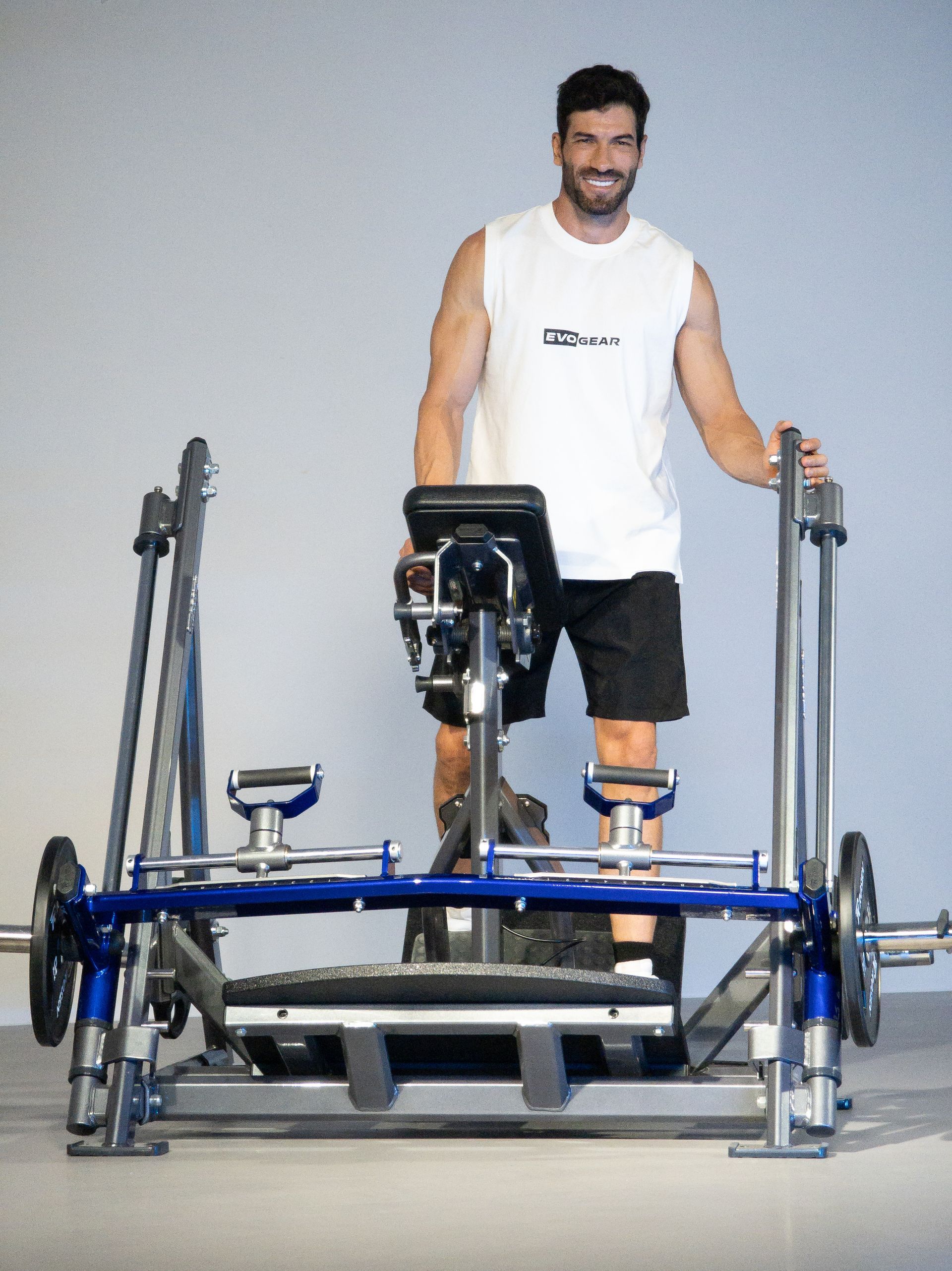 Homem em pé em um aparelho de ginástica azul, segurando as barras e sorrindo em um estúdio de fitness.