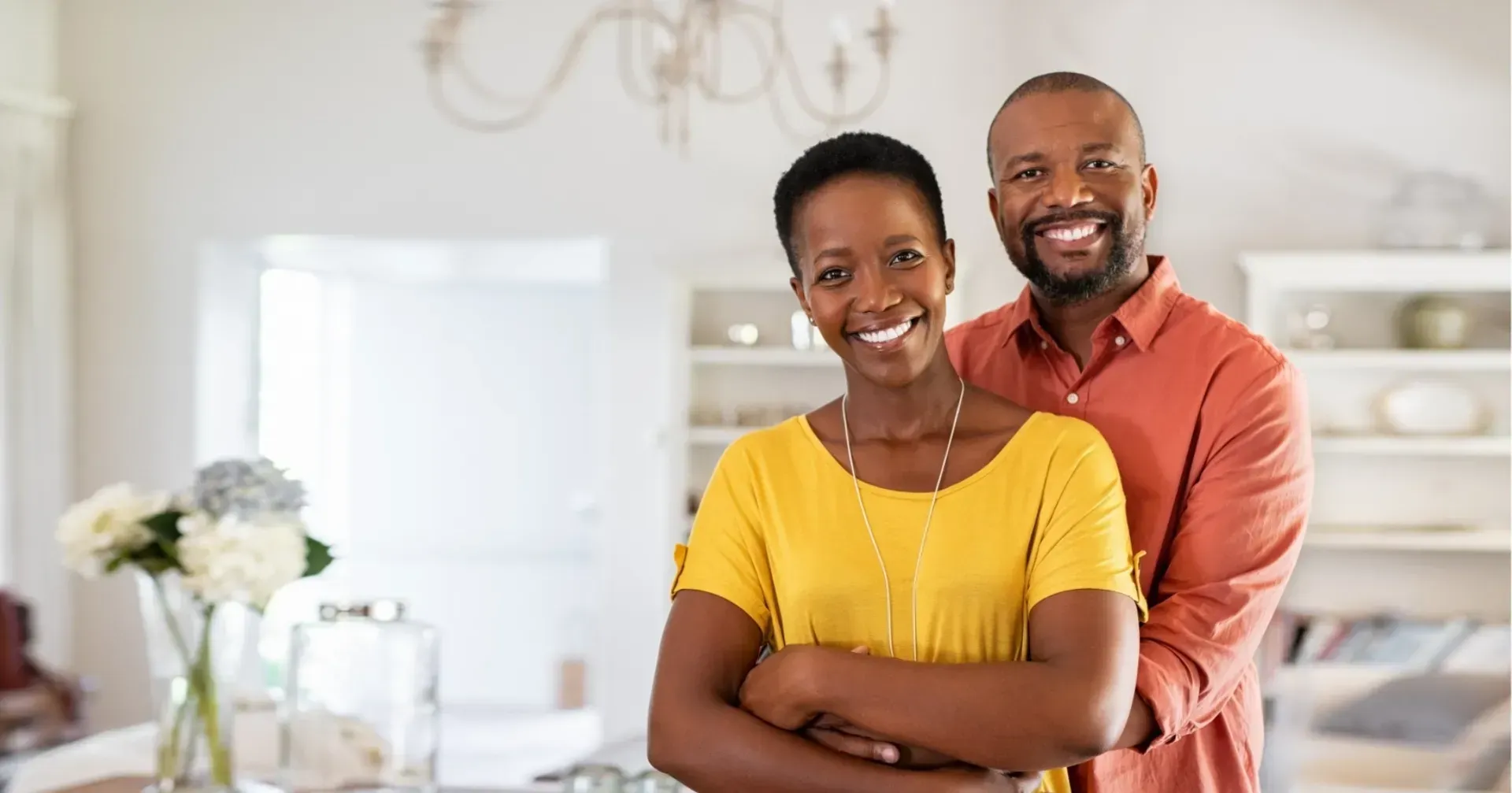 A smiling couple poses for a portrait in a bright, modern kitchen with white cabinetry and a vase of flowers.