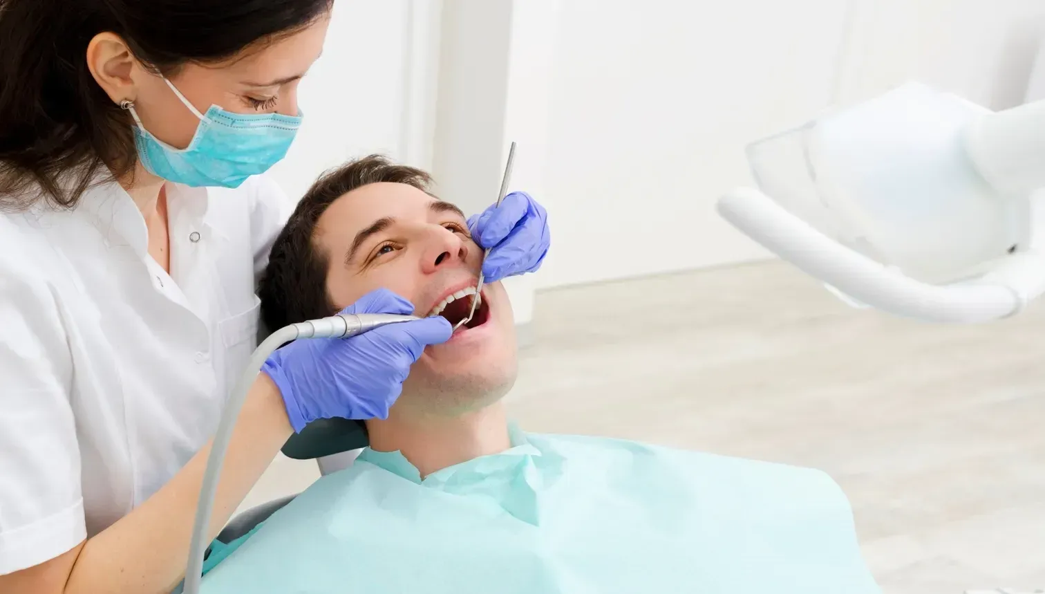 A dental professional in a white coat and blue mask examines a patient's teeth with a tool in a clinical setting.