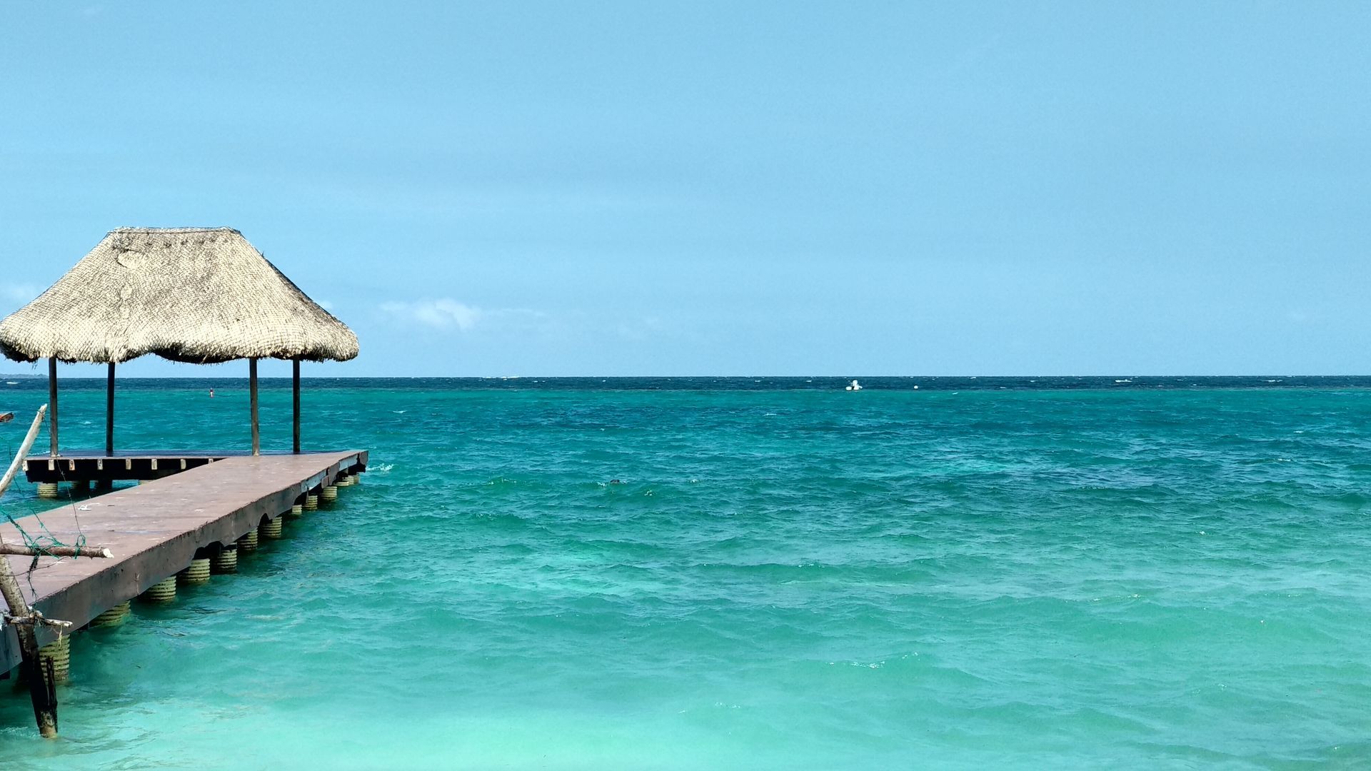 Wooden pier with thatched roof shelter over turquoise ocean under a clear blue sky.