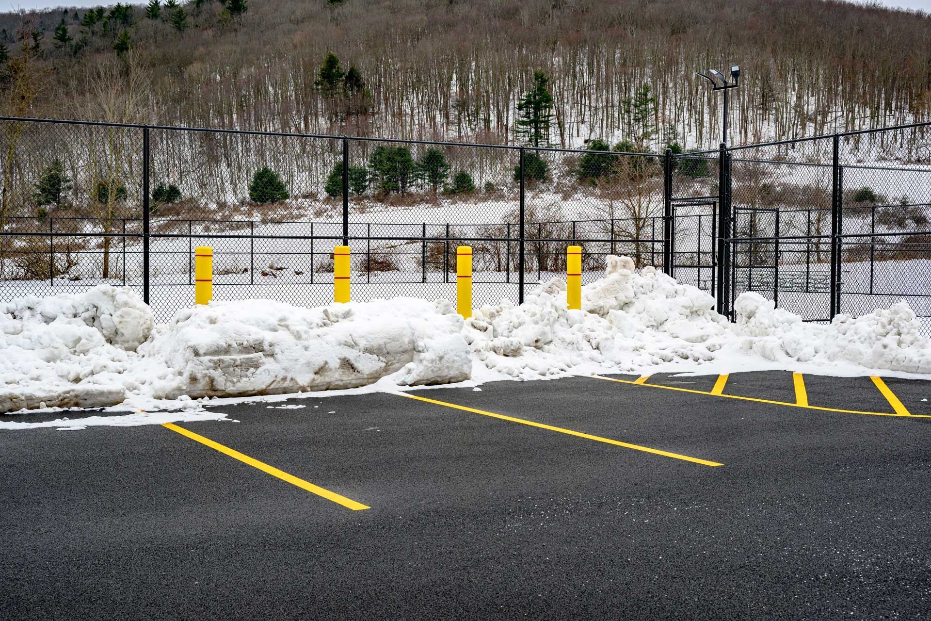 Snow-covered parking lot with yellow painted lines and bollards; a chain-link fence and a snowy forest in the background. Snow-covered parking lot with yellow painted lines and bollards; a chain-link fence and a snowy forest in the background.