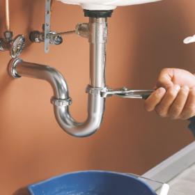 Un plombier utilise une clé sur le tuyau d'évacuation d'un évier. Un bol bleu recueille l'eau en dessous.