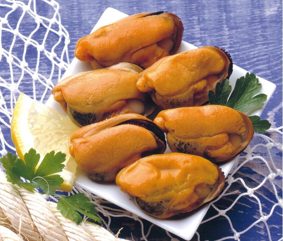 Una ración de seis mejillones grandes y anaranjados en un plato blanco, adornados con una rodaja de limón y perejil fresco sobre una superficie azul.