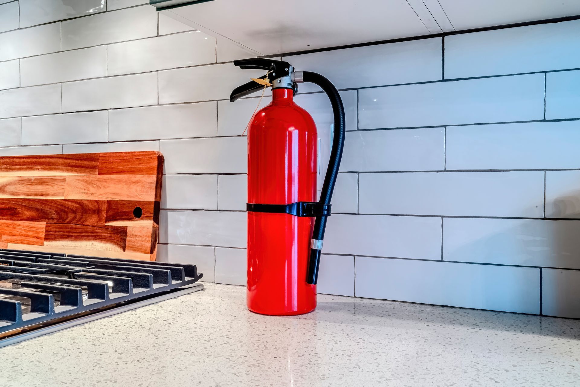 Red fire extinguisher on countertop in a kitchen with white tile backsplash and wooden cutting board.