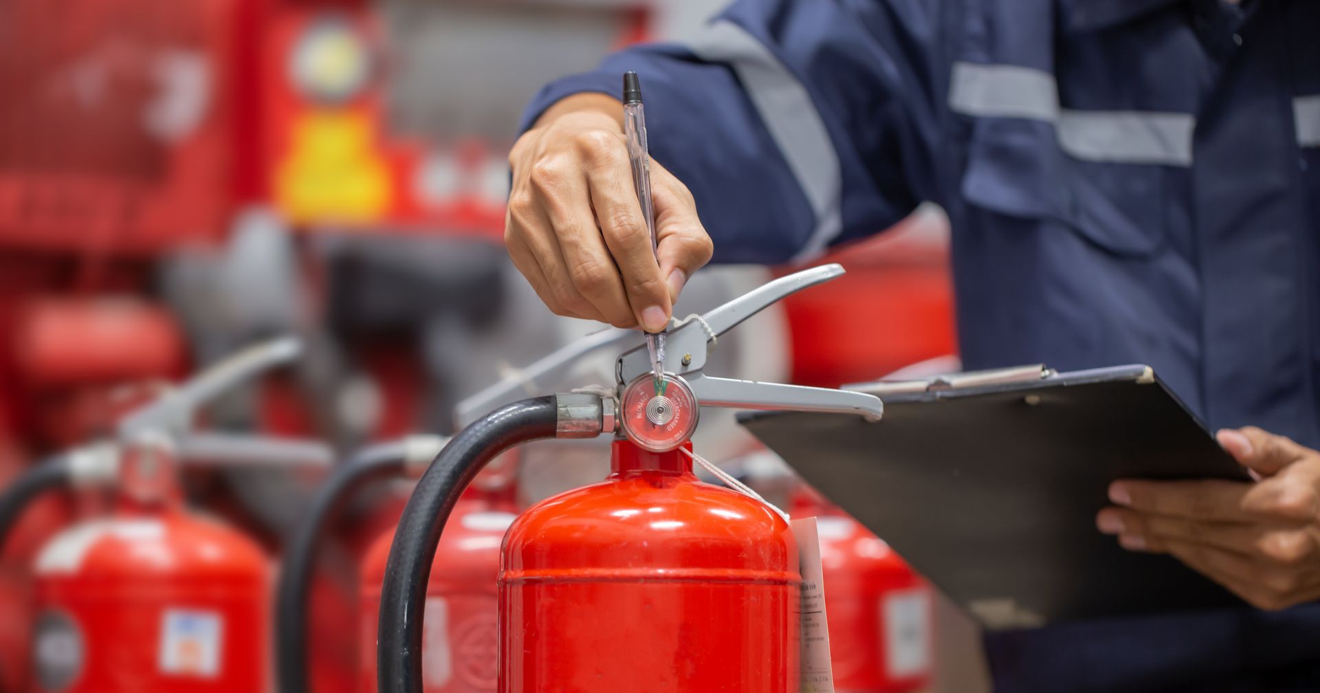 Person in blue uniform inspecting a red fire extinguisher, holding a clipboard, in a room with other fire extinguishers.