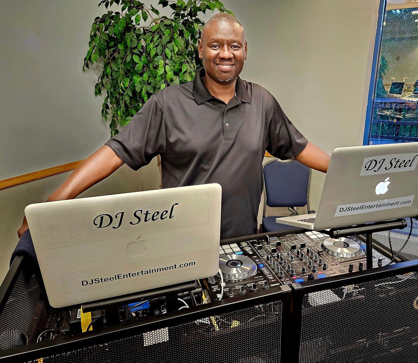 Man standing behind a DJ booth with two laptops, smiling in a room with a wall painting.