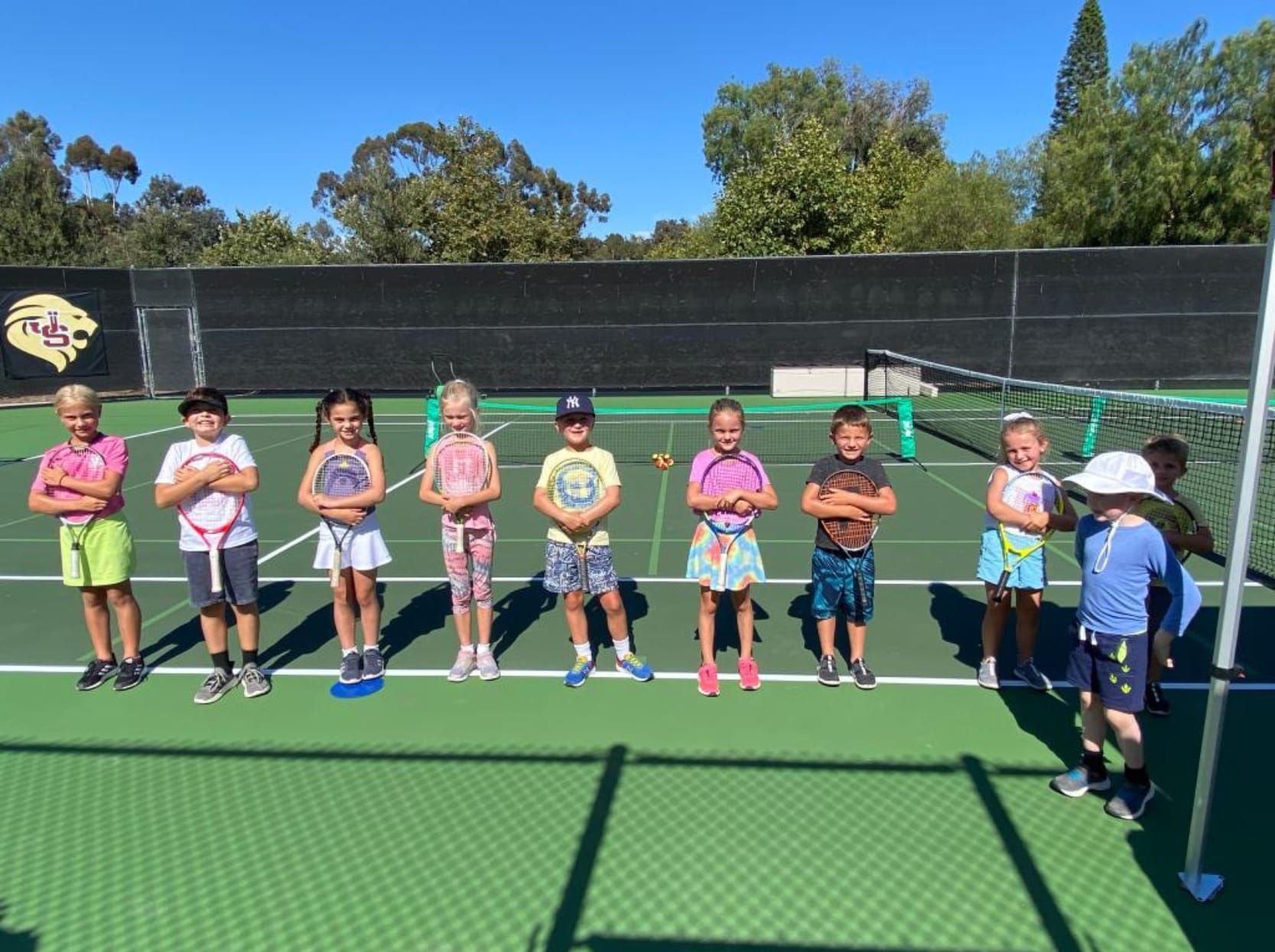 A group of children are standing on a tennis court holding tennis rackets.