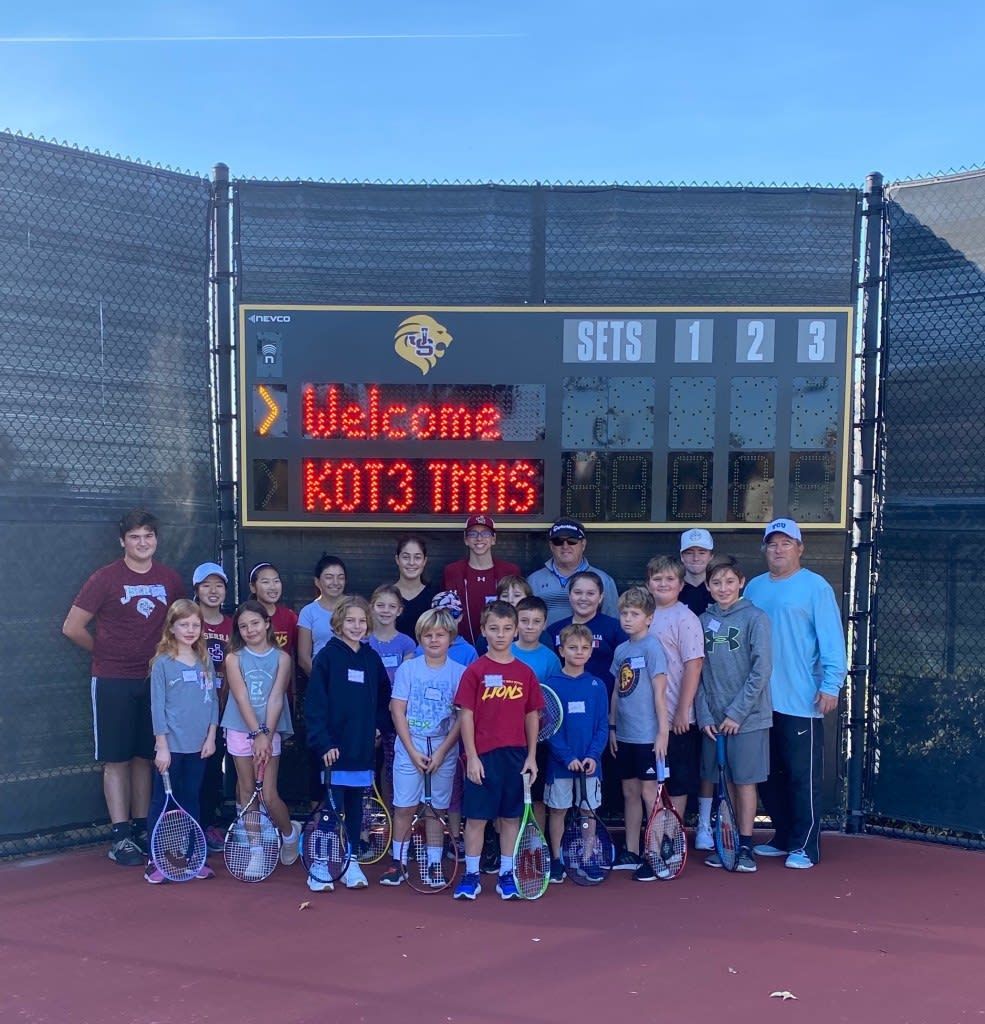 A group of kids standing in front of a scoreboard that says welcome kot3 tennis