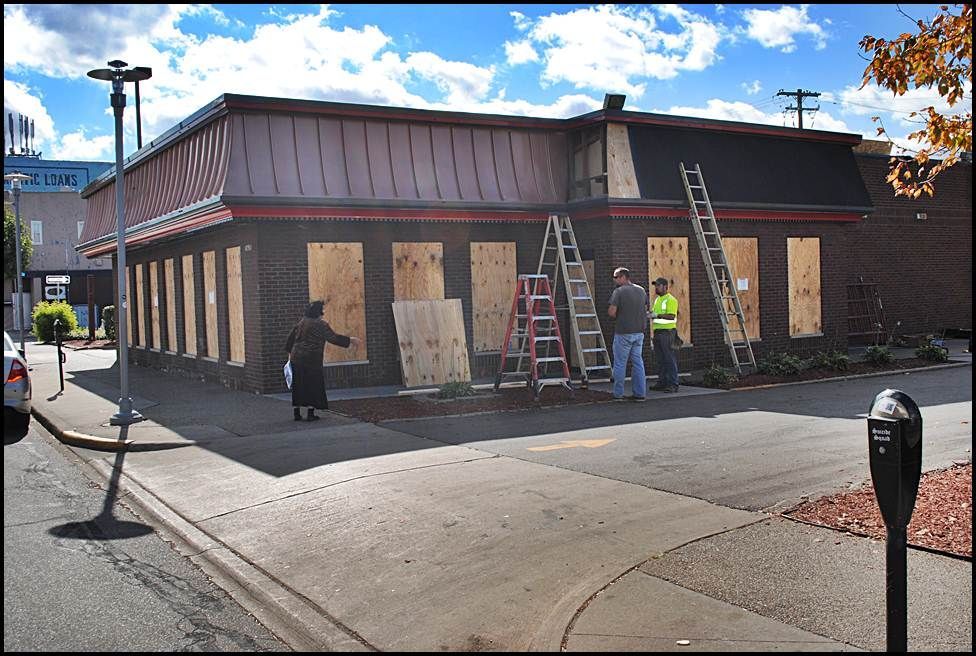 Building under construction, boarded windows, workers near ladders, sunny day, street view.