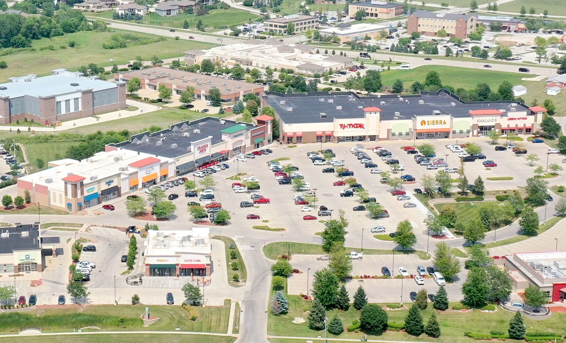 Aerial view of a shopping center with various stores, parking lot filled with cars, and green surroundings.