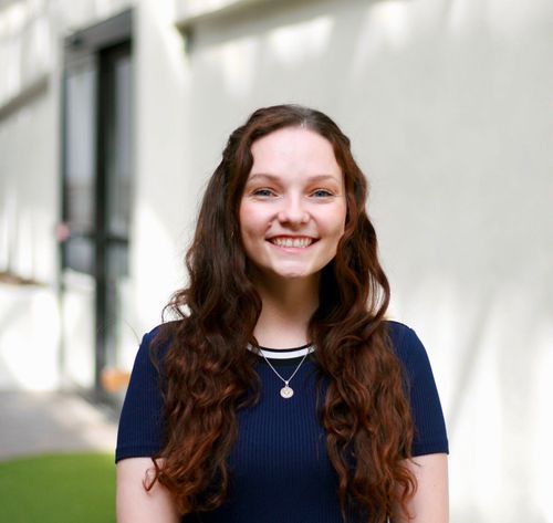 Smiling person with long wavy hair wearing a navy shirt and necklace, standing outdoors by a light wall.