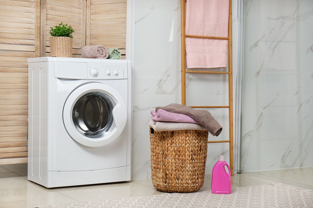 White Washing Machine Stands Next to a Wicker Laundry Basket — Eurocoast Appliance Service In Gosford, NSW