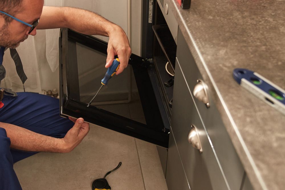 Person Repairs an Open Oven Door Using a Screwdriver in a Kitchen — Eurocoast Appliance Service In Woy Woy, NSW