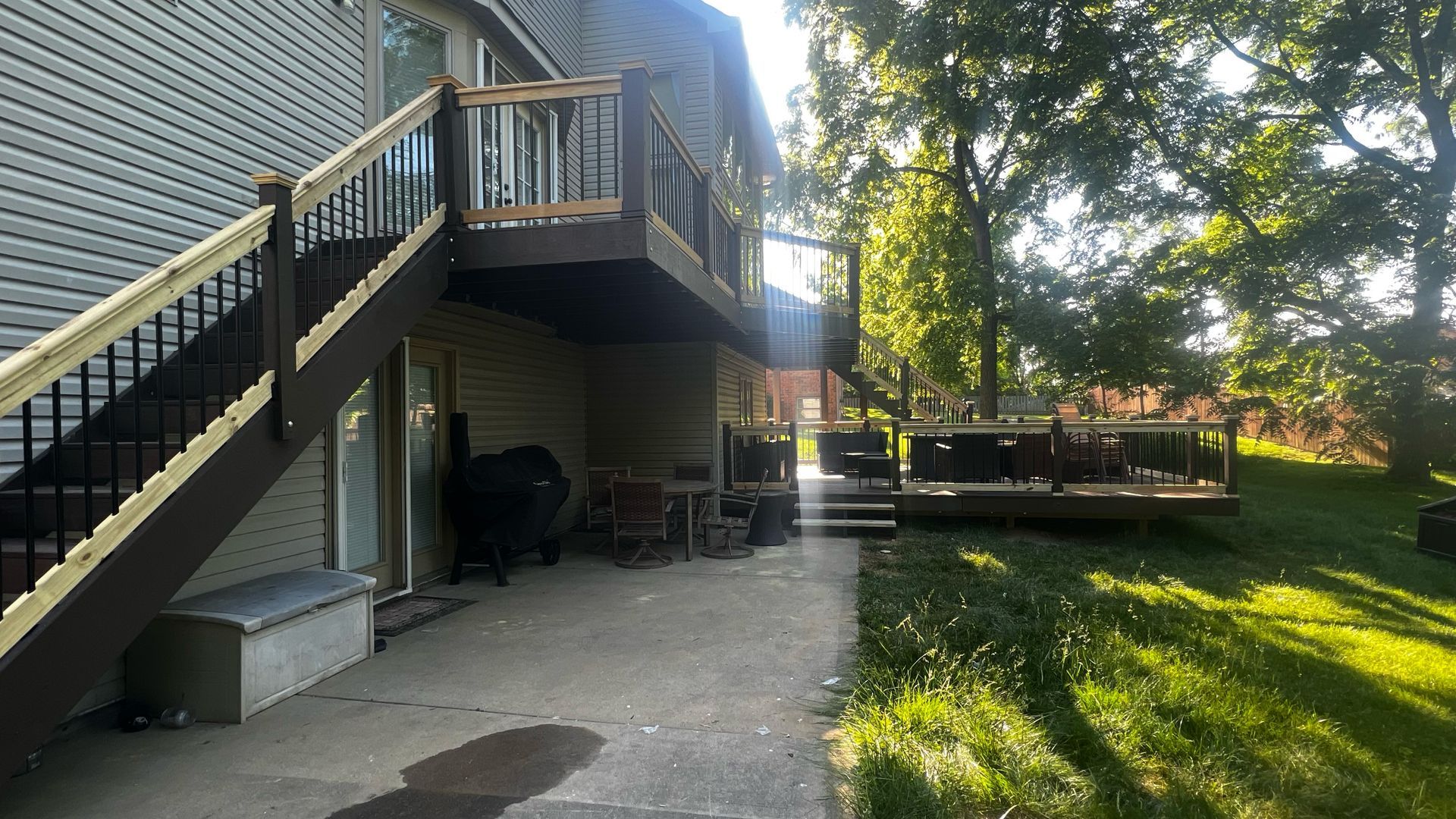 Two-story house with decks, stairs, and a concrete patio. Trees and grass surround the backyard.