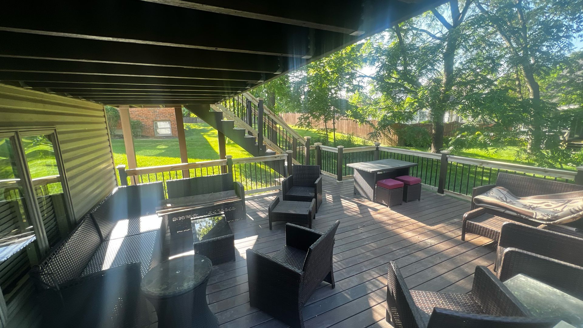 Outdoor deck with black furniture, beneath a wooden structure, overlooking a yard with trees.