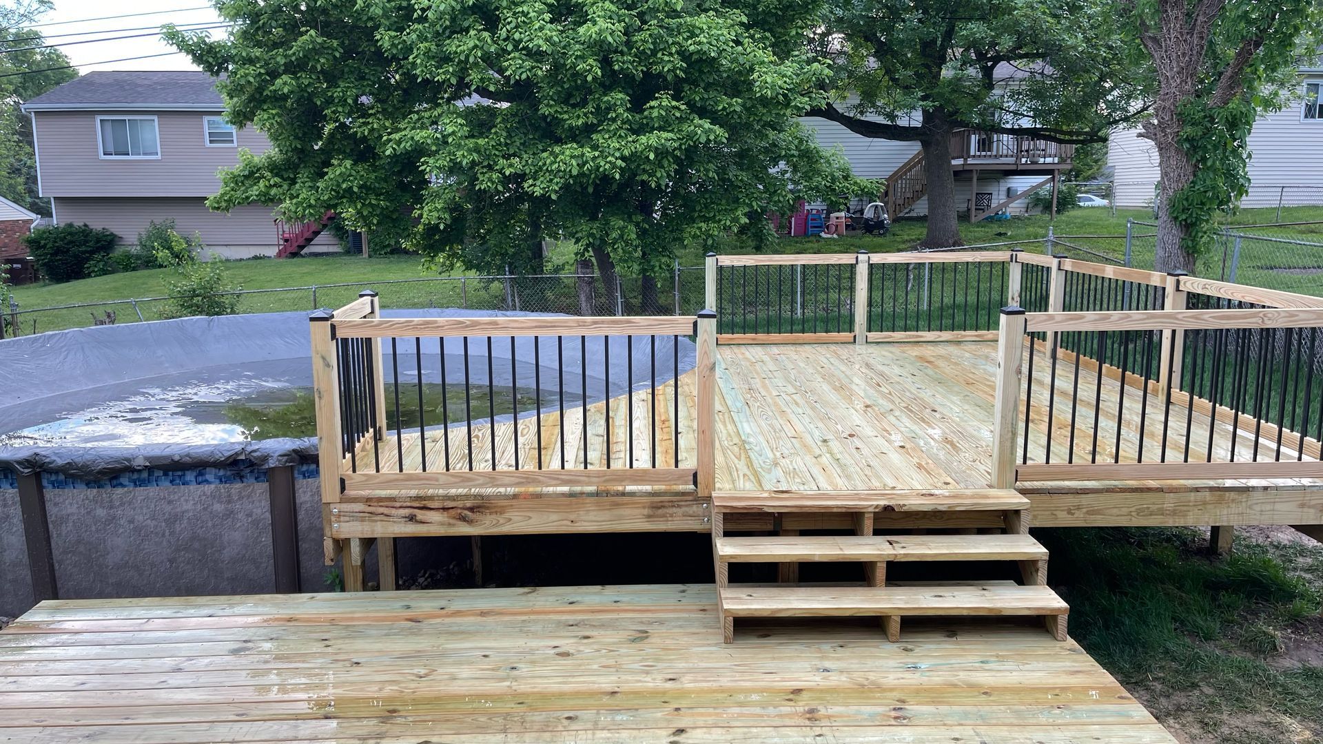 Wooden deck with steps and black railing next to an above-ground pool and grass lawn.