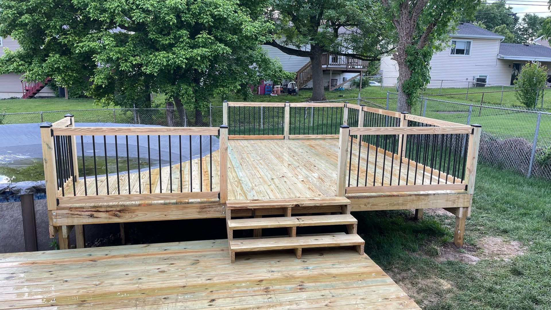 Wooden deck with steps and black railing; outdoors with grass, trees, and houses in the background.