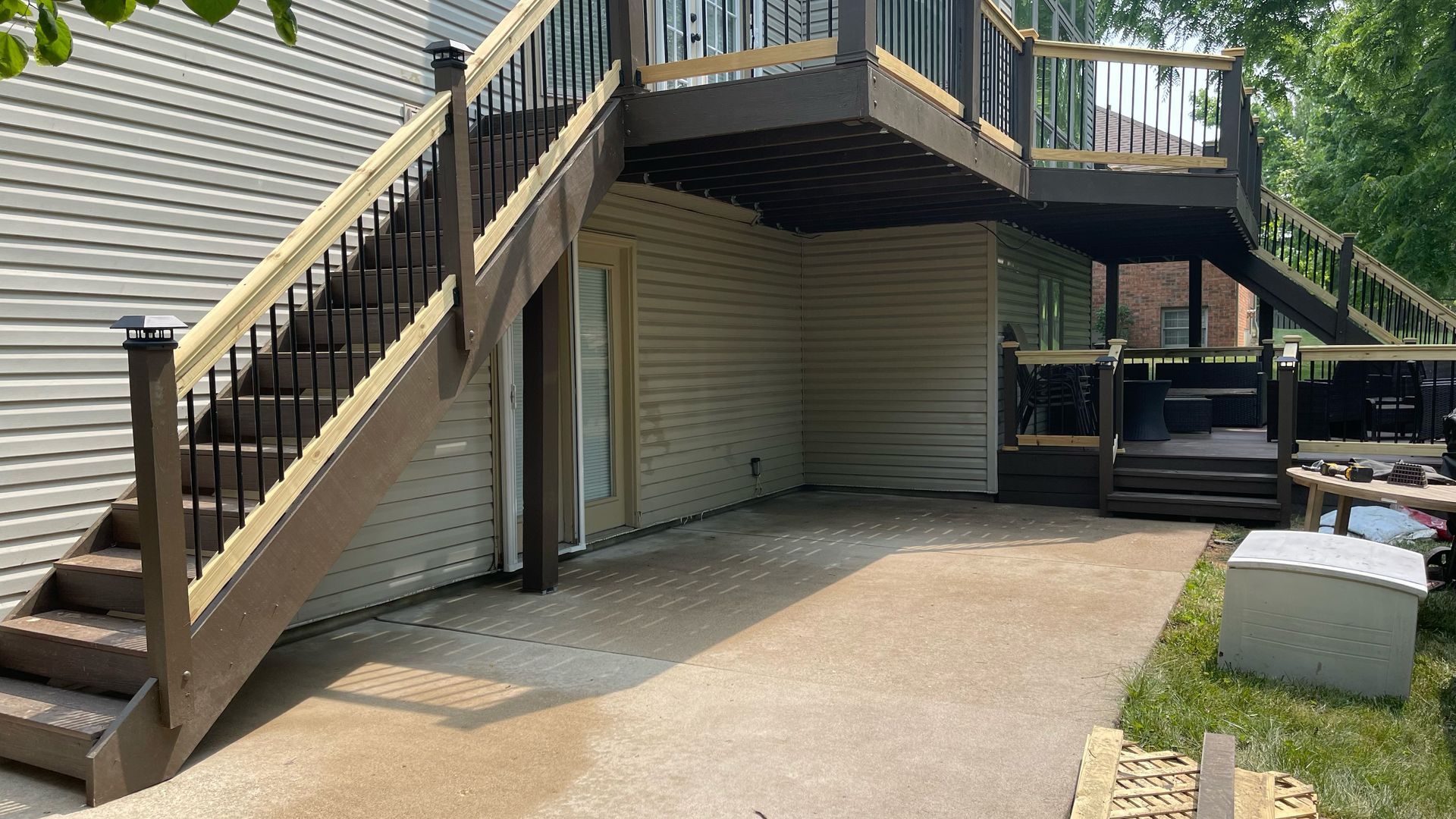 Two-story deck with stairs leading down to a concrete patio. Beige siding, brown deck and railing, black balusters.