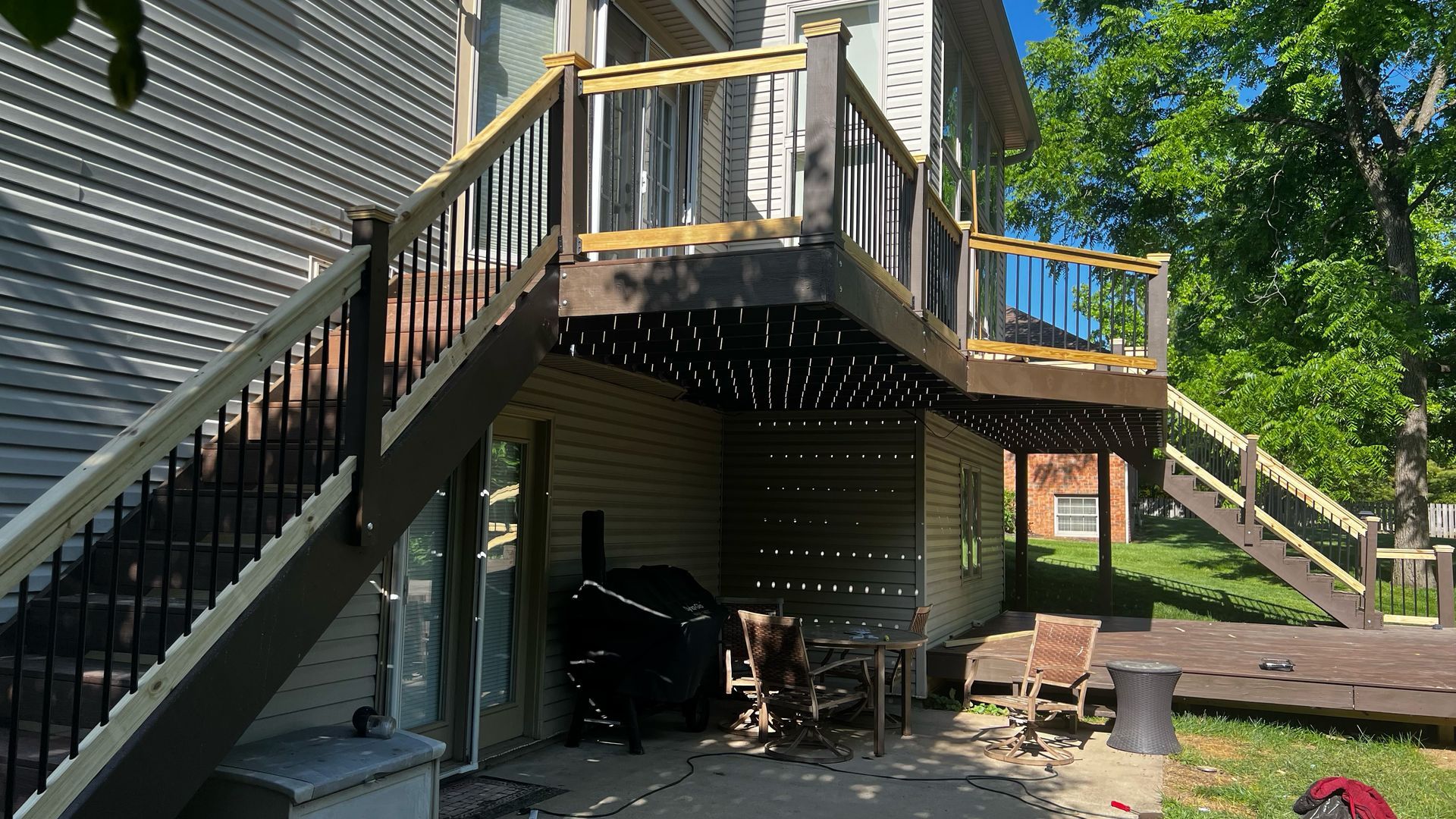 Two-story wooden deck with stairs. Beige siding on the house. Black and brown railing. Outdoor seating underneath.