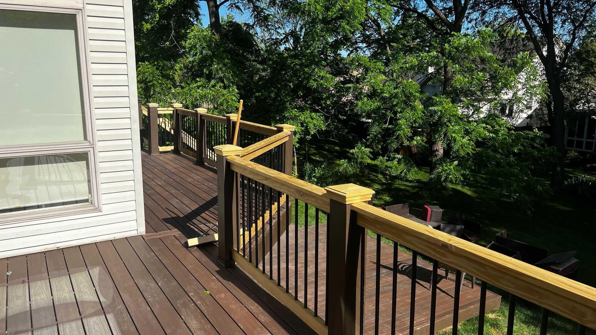 Wooden deck with black railing and light brown trim, overlooking lush green trees.