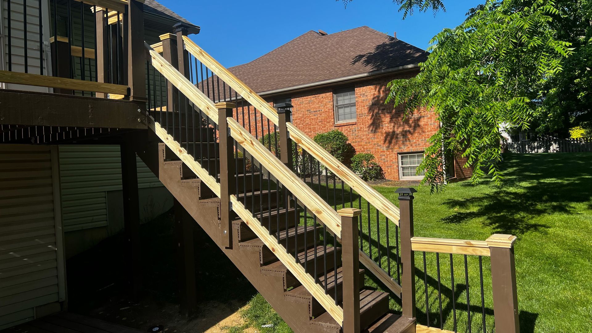 Wooden deck stairs with railings leading down, brown and black tones, in a backyard setting.