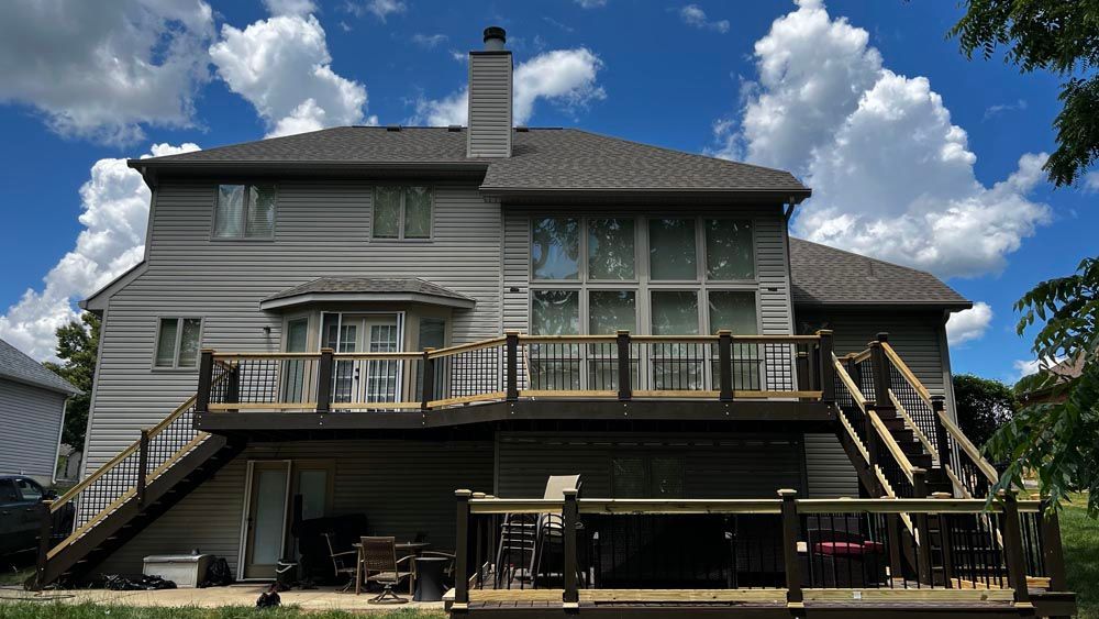 Two-story gray house with a large deck, stairs, and a cloudy blue sky above