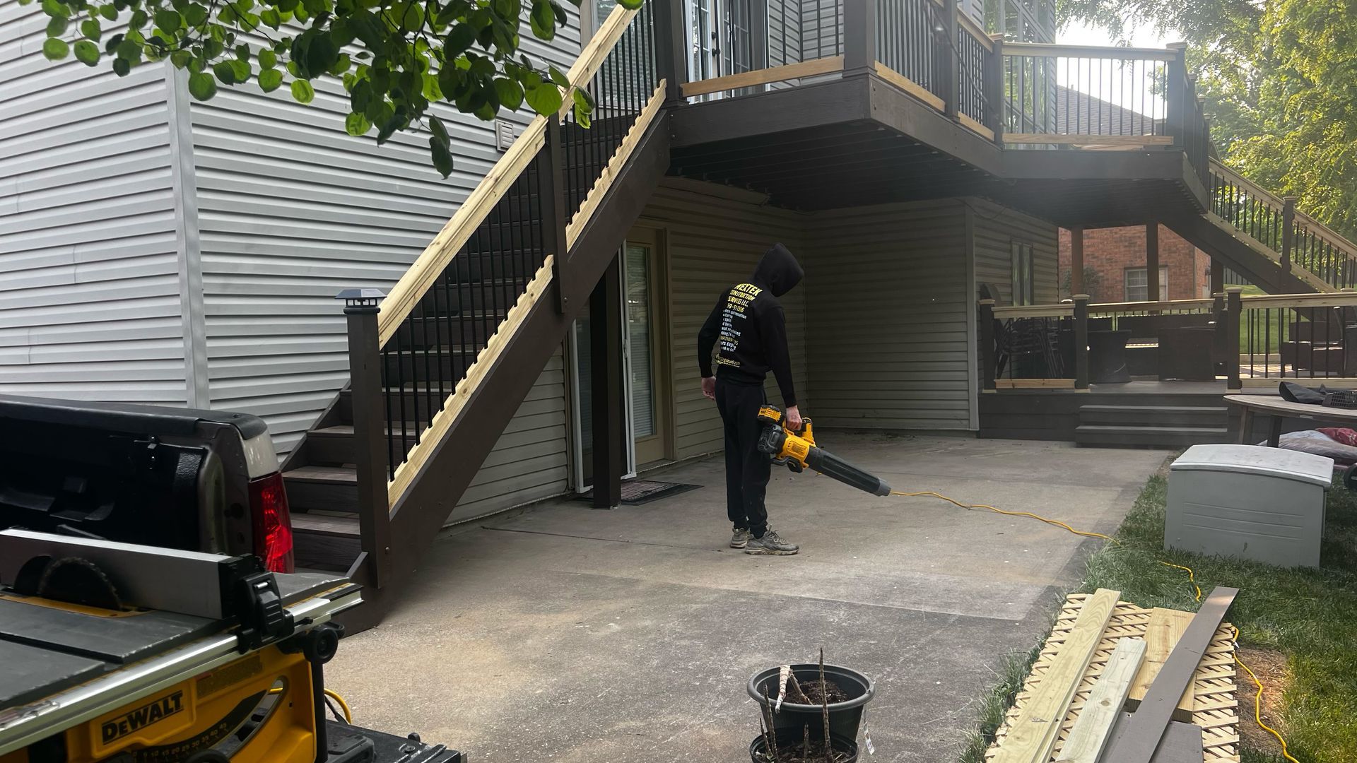 A person using a leaf blower on a concrete patio next to a deck with stairs. A truck and tools are nearby.
